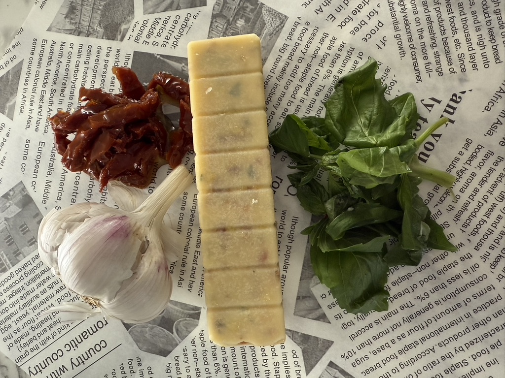A portioned flavoured butter beside sun-dried tomatoes, garlic and fresh basil.